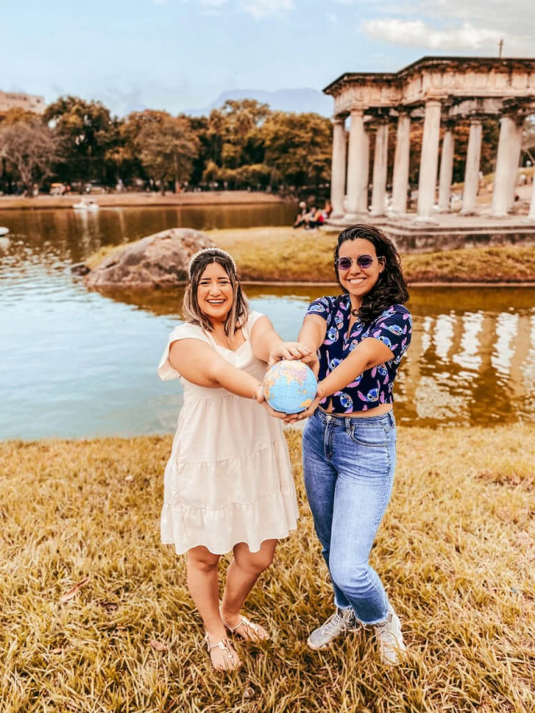 Foto de duas moças com os braços estendidos à frente de seu corpo segurando um globo, a moça do lado esquerdo tem a pele clara e veste um vestido bege, ela sorri. A moça do lado direito também tem a pele clara, ela veste uma blusa azul do Little Stitch e calça jeans e também usa um óculos de sol. Ao fundo, há um lago e uma construção greco-romana