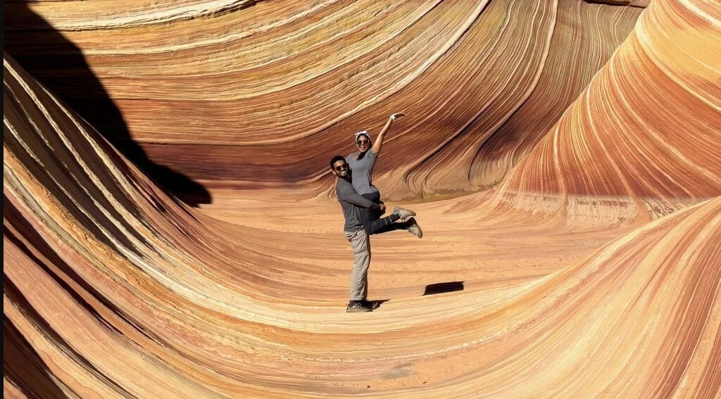 um homem levanta uma mulher no meio do deserto enquanto ambos olham para a câmera e sorriem